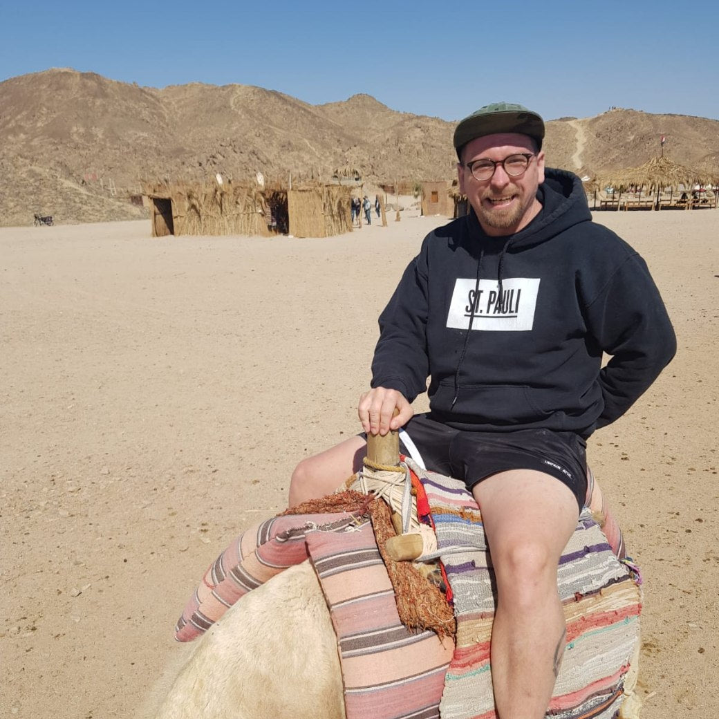 Originalfoto: Mann mit Mütze und Kapuzenpulli auf einem Kamel in der Wüste